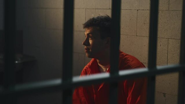 Prisoner in orange uniform sits in prison cell. Cellmate criminal speaks aggressively, threatens him. Inmates serve imprisonment terms for crimes in jail. Detention center or correctional facility.