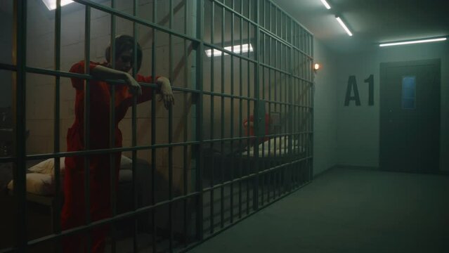 One female prisoner in orange uniform stands behind metal bars, another sits on the bed in prison cell. Women serve imprisonment terms for crimes in jail. Depressed inmates in correctional facility.