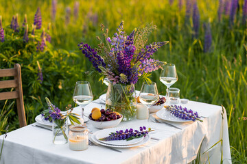 Beautiful romantic outdoor wedding decor in a field. Table decorated with purple lupines flowers. Wineglasses with white wine. Sunset, summer, golden hour. Perfect surprise date for loving couple
