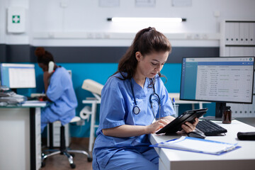 Young nurse checking appointments list on digital tablet in busy medical office. Adult woman healthcare specialist working with technology at hospital desk with stethoscope around neck - Powered by Adobe