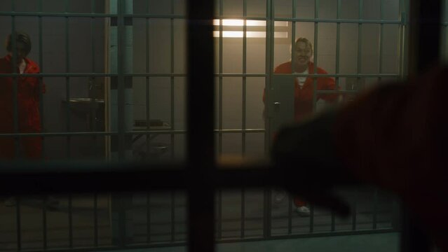 Insane female prisoner in orange uniform yells at neighbor inmate, jumps on metal bars in prison cell. Women serve imprisonment terms in jail. Criminals in correctional facility. Justice system.