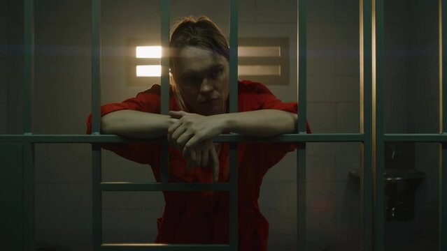 Female prisoner in orange uniform holds hands on metal bars, walks, looks at barred window in jail cell. Woman serves imprisonment term for crime in prison. Depressed inmate in correctional facility.