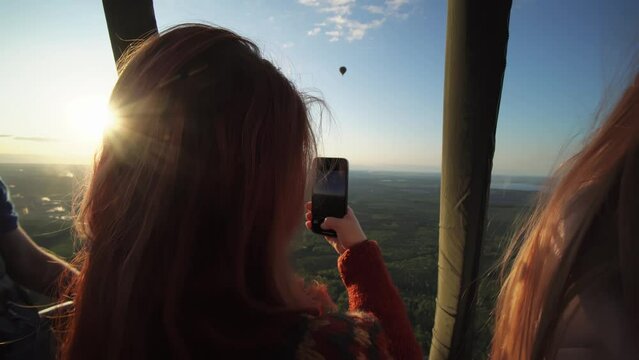 Woman is taking picture on smartphone in her hands flying inside basket of hot air balloon. Caucasian cheerful lady is enjoying picturesque sunset being on excursion with her friends. Active weekend