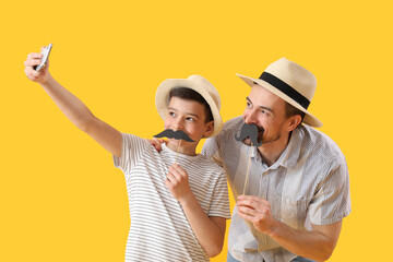 Portrait of father and his little son with paper mustache taking selfie on yellow background