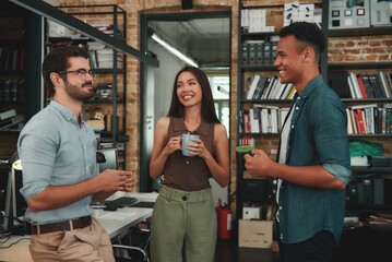 Fresh office news. Young smiling colleagues in casual wear holding cups and talking about something while standing in modern office