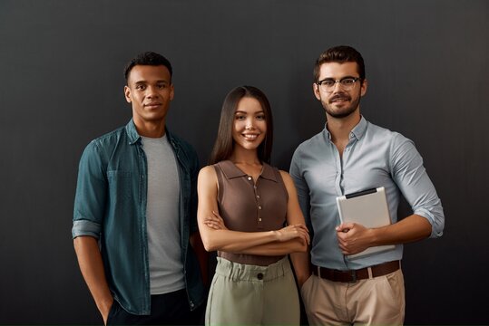 Perfect IT team. Group of three cheerful young people in casual wear looking at camera with smile while standing against dark background