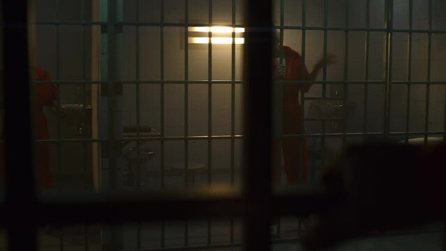 One angry female prisoner in orange uniform yells at neighbor inmate and jumps on bars, another nervously walks in prison cell. Women serve imprisonment terms for crimes in jail. Detention center.