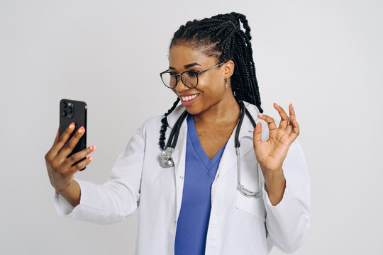 Portrait Of Happy African American Nurse Or Doctor Woman Wearing Medical Lab Coat Holding And Using Cellphone Isolated Against White Background