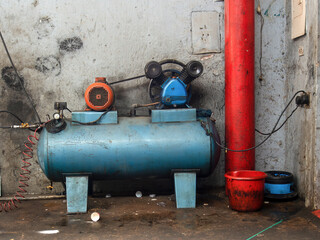 A rustic and very used blue air compressor at the side of a red column in a dirty car shop in the town of Arcabuco, in central Colombia.