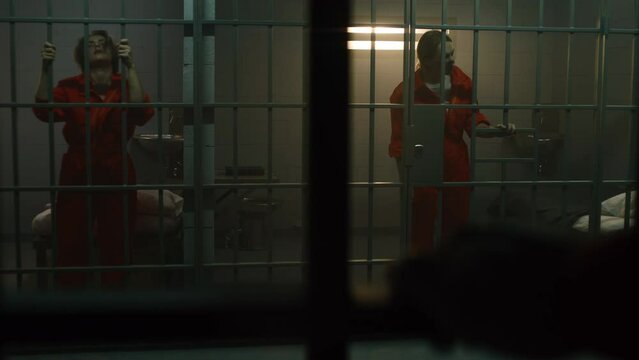 Two female prisoners in orange uniforms stand and walk behind bars in jail cell. Criminals serve imprisonment terms for crimes in prison. Women inmates in detention center or correctional facility.