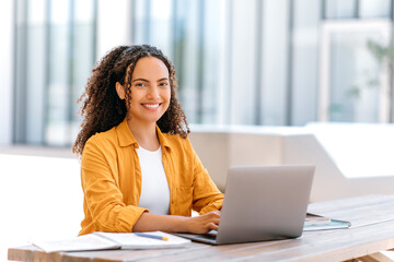 Fototapeta na wymiar Photo of a successful curly haired girl, brazilian or hispanic nationality, outsourcing employee or student, uses laptop to work or study online while sitting outdoors, looks at camera, smile friendly
