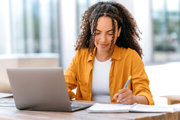 Education concept. Positive hispanic or brazilian curly haired girl, student or freelancer, sit outdoors with a laptop, working or studying online, writing down information in her notepad, smiles