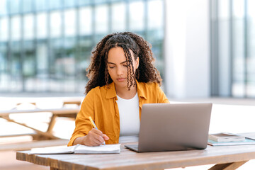 Focused trendy hispanic or brazilian curly haired girl, student or freelancer, sitting outdoors with a laptop, working or studying online, concentrated writing down information in her notepad - obrazy, fototapety, plakaty
