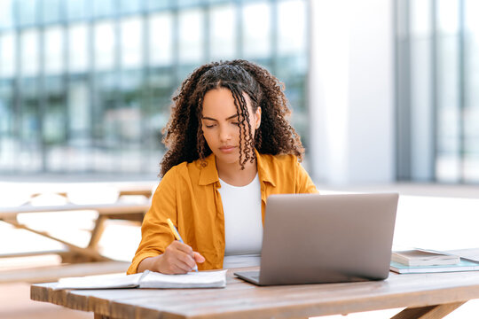 Focused trendy hispanic or brazilian curly haired girl, student or freelancer, sitting outdoors with a laptop, working or studying online, concentrated writing down information in her notepad