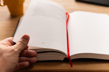 Close-up of a hand turning page of book with a cup of tea next to it on wooden table