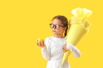 Happy little girl in eyeglasses with school cone and fresh apple on yellow background