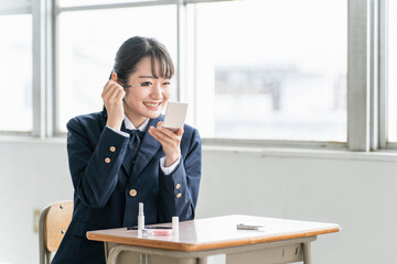 放課後に学校の教室で鏡を見ながらメイクをする笑顔の女子高生（マスカラ・アイメイク）
