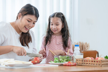 Asian mother and daughter preparing breakfast in kitchen, cute child looking at mom cutting apples, making salad and sandwich, milk, bread on table. Happy family, healthy food at home. Focus on kid