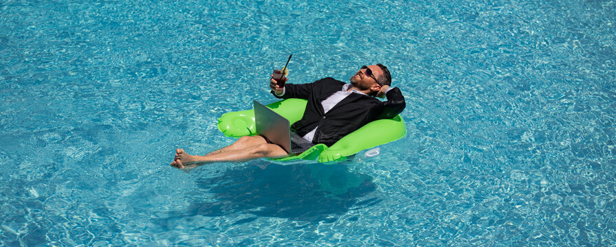 Funny business man in a business suit floating in the water in the pool. Remote summer work online. Crazy freelancer. Business and summer. Business man drink summer cocktail and using laptop in pool. - Powered by Adobe