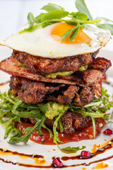 A stacked breakfast dish with fried egg, bacon, vegetable fritters, avocado and tomato relish, served on a white round plate.