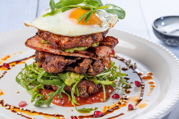 A stacked breakfast dish with fried egg, bacon, vegetable fritters, avocado and tomato relish, served on a white round plate.