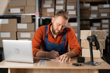 Inventory manager small business owner working on laptop in warehouse, writing delivery address on...