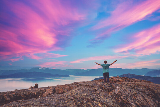 Panoramic view of the fjord. Twilight with a pink sky. A young man with hands in the air standing on a cliff of rock