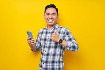 Smiling satisfied young Asian man wearing a white checkered shirt holding a mobile phone and showing thumbs up sign, recommends apps isolated over yellow background. People Lifestyle Concept