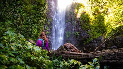 adventurous hiker girl standing in front of large tropical waterfall surrounded by lush green...