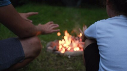 Selective focus of young dad and daughter sitting near bonfire on background forest at night. Little girl with father enjoys summer nature. Outdoor camp vibes lifestyle, living outside urban nomad