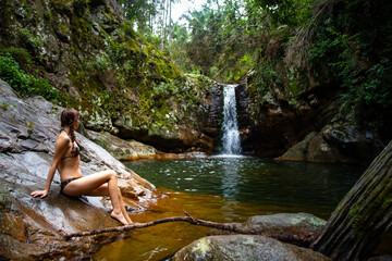 beautiful long hair woman in bikini sitting on the edge of a rock pool in front of large tropical waterfall at mount barney national park, queensland, australia	