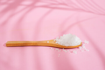 Wooden tea spoon filled with white rock salt, on pink background with palm leaf shadows,