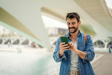 A happy hipster is standing in urban exterior and typing on the phone.