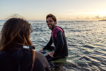 Fototapeta premium Happy surfers laughing at each other while sitting on the surfboards in water.