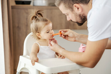 A father is giving medicine to his ill daughter trough the syringe while she is sitting in chair.