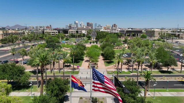 American flag, Arizona state flag, and POW MIA flags waving in on capitol grounds. Aerial rising shot revealing Phoenix, Arizona skyline.