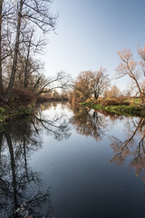 Fototapeta na wymiar River with trees around mirroring on water ground during autumn day with clear sky