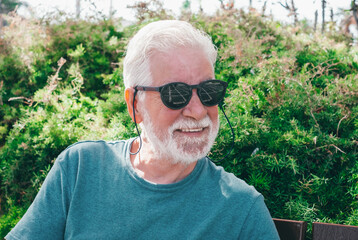 Naklejka na ściany i meble Close up portrait of happy senior bearded man sitting in the park on a bench. Elderly smiling grandfather with black sunglasses relaxes enjoying vacation or retirement