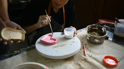 Young creative asian man painting clay plate with a paintbrush at desk in ceramic workshop