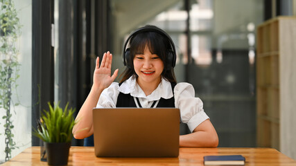 Cute Asian young woman in wireless headphone studying remotely, listening to online lecture on laptop