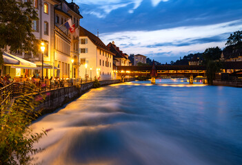 Fototapeta na wymiar Night time panorama in Lucerne old town in Switzerland. Colorfully illuminated waterfront of river Reuss with strong current in evening blue hour twilight. Wooden bridge, cafes and city wall tower.