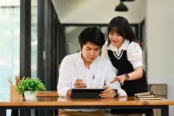 Young female office worker pointing at digital tablet screen, discussing on new project with colleague