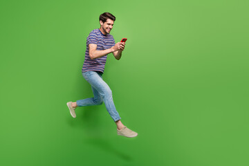 Full length photo of cute positive man dressed striped t-shirt jumping high texting modern device empty space isolated green color background