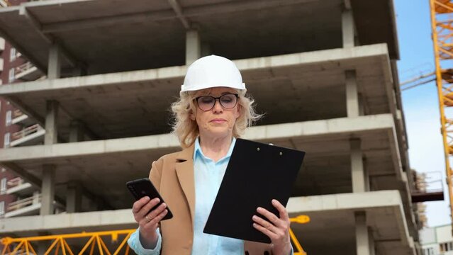 Senior woman entrepreneur in protective helmet types data to client on phone from clipboard on background of building. Concept of construction and working moments