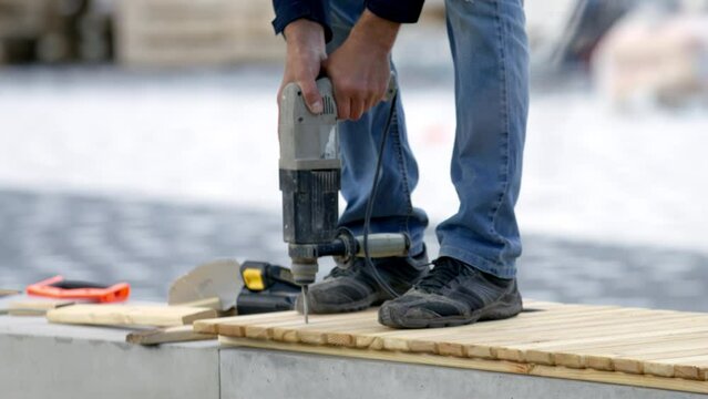 Person in ripped jeans drills wooden blanks and stands on the bench at daytime. Worker attaches boards of wood together to create bench during park renovation