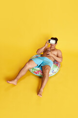 Full-length image of young man in swimming trunks and vr glasses, sitting into swimming circle against yellow studio background. Concept of summer, vacation, leisure time, holidays, human emotions, ad