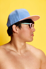 Profile portrait of young man in sunglasses and cap posing shirtless against yellow studio background. Tanned body. Concept of summer, vacation, leisure time, holidays, human emotions, ad