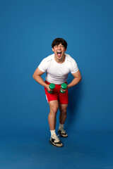 Fototapeta na wymiar Full-length portrait of young man in sportswear training with dumbbells and having fun against blue studio background. Concept of sport, fitness, active and healthy lifestyle, human emotions, ad