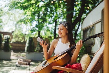 Happy hippie girl is having a good time with playing on guitar with smartphone in her hand in camper trailer. Holiday, vacation, trip concept.