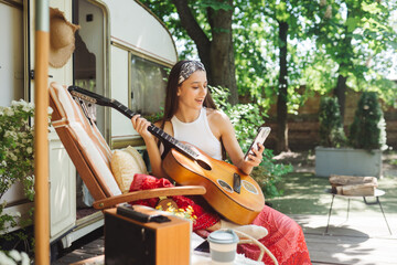 Happy hippie girl is having a good time with playing on guitar with smartphone in her hand in...